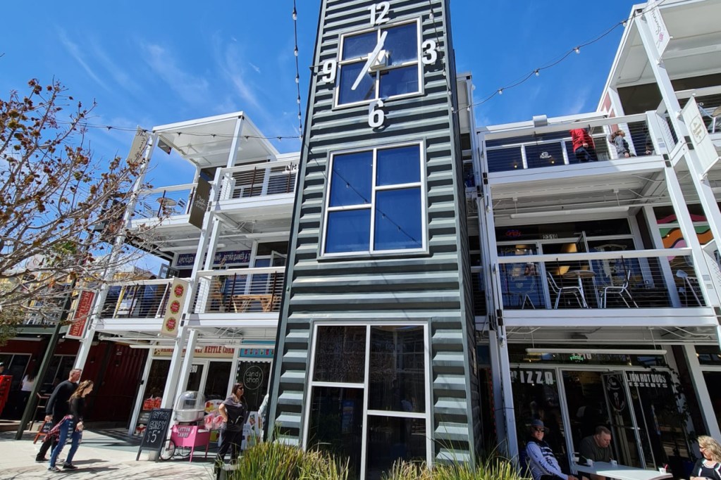 Shipping Container Clock Tower and Commercial Complex 2 A multi-level commercial building made from shipping containers, featuring a clock tower structure in the center, with outdoor seating and shops at the base.